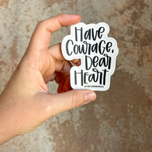 Load image into Gallery viewer, A white woman holds up a sticker containing black text "Have Courage, Dear Heart" with a white background color in front of a rustic brown wall.