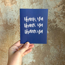 Load image into Gallery viewer, A dark blue card with white text "thank ya" printed three times on three separate lines. The card is held in front of a rustic brown wall by a white woman.