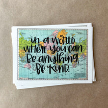 Load image into Gallery viewer, Stack of horizontal sheets of paper; paper on top is a blue, green, yellow, and pink flat map of the world with text "in a world where you can be anything, be kind" in black lettering over the map print. The paper is lying on a large piece of brown paper.