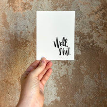 Load image into Gallery viewer, A white card with black text "Well, shit." in the bottom right corner. The card is held in front of a brown rustic wall by a white woman.