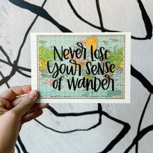 Load image into Gallery viewer, A blue, green, yellow, and pink flat map of the world with text "never lose your sense of wonder" in black lettering over the map print. The map is held in front of a white backdrop with spiraling black lines by a white woman.