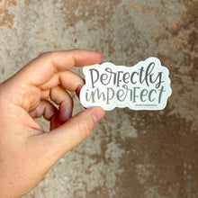 Load image into Gallery viewer, Text "perfectly imperfect" in two different colors. "Perfectly" is gray, and "imperfect" is green. The background color is white. The sticker is held by a white woman, in front of a brown rustic wall.