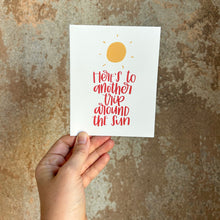 Load image into Gallery viewer, A white card with red text "Here's to another trip around the sun" below a stylized yellow sun design with short lines radiating outwards to depict rays of sunlight. The card is held in front of a brown rustic wall by a white woman.