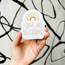 Load image into Gallery viewer, Light pink text "you are worthy" under a 4 stripe rainbow. The inner stripe is a golden brown. The next stripe is light pink with white arrow tip designs. The third stripe is a dashed golden line. The outer stripe is a blue-green line with a thin white line in the middle of it. The sticker is held in front of a white backdrop with spiraling black lines by a white woman.