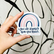 Load image into Gallery viewer, Dark blue text "You are stronger than you think" under a four stripe rainbow. The inner stripe is thin and dark blue, the next stripe is thick and light pink with a thin pink spiral inside, the third stripe is thin and pink, and the last stripe is slightly thicker and dark blue. The background color is white. The sticker is held in front of a white backdrop with black spiraling lines by a white woman.
