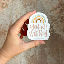 Load image into Gallery viewer, Light pink text "you are worthy" under a 4 stripe rainbow. The inner stripe is a golden brown. The next stripe is light pink with white arrow tip designs. The third stripe is a dashed golden line. The outer stripe is a blue-green line with a thin white line in the middle of it. The sticker is held in front of a brown rustic wall by a white woman.
