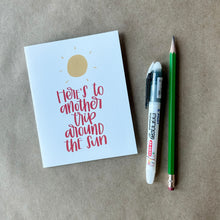 Load image into Gallery viewer, A white card with red text "Here's to another trip around the sun" below a stylized yellow sun design with short lines radiating outwards to depict rays of sunlight. The card is lying on a brown piece of paper beside a pencil and a pen.