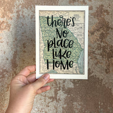 Load image into Gallery viewer, A vintage style map design of the state of Georgia with black lettered text "there's no place like home" over the print. The map is held in front of a brown rustic wall by a white woman.