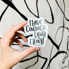 Load image into Gallery viewer, A white woman holds up a sticker containing black text "Have Courage, Dear Heart" with a white background color in front of a white wall with a spiraling black stripe design.