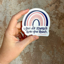 Load image into Gallery viewer, Dark blue text "You are stronger than you think" under a four stripe rainbow. The inner stripe is thin and dark blue, the next stripe is thick and light pink with a thin pink spiral inside, the third stripe is thin and pink, and the last stripe is slightly thicker and dark blue. The background color is white. The sticker is held in front of a brown, rustic wall by a white woman.