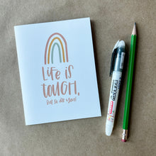 Load image into Gallery viewer, A white card with orange text "Life is tough, but so are you!" under a three stripe rainbow. The inner stripe of the rainbow is a green line, the middle stripe is an orange line, and the outer stripe is a red line. The card is lying on a brown piece of paper beside a pencil and a pen.