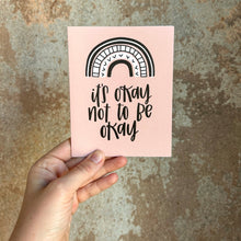 Load image into Gallery viewer, A light pink card with black text "it's okay to not be okay" under a four stripe rainbow. The inner stripe is a thin black line. The next stripe is a white line with arrowhead designs. The third stripe is a white line with thin black lines sectioning it into pieces. The outer stripe is a black line with a thinner white line inside it. The card is held in front of a brown rustic wall by a white woman.