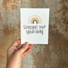 Load image into Gallery viewer, A white card with gray text "Sending love your way" under a four stripe rainbow. The inner stripe is a dark gray line. The next stripe is a golden line. The third stripe is a lighter gray line. The outer stripe is a line of red dots. The card is held in front of a brown rustic wall by a white woman.