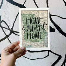 Load image into Gallery viewer, A vintage style map design of the state of Georgia with black lettered text "Home Sweet Home" over the print. The map is held in front of a white backdrop with spiraling black lines by a white woman.