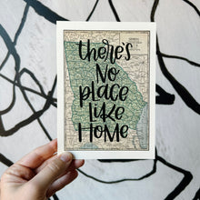 Load image into Gallery viewer, A vintage style map design of the state of Georgia with black lettered text "there's no place like home" over the print. The map is held in front of a white backdrop with spiraling black lines by a white woman.