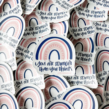 Load image into Gallery viewer, Dark blue text "You are stronger than you think" under a four stripe rainbow. The inner stripe is thin and dark blue, the next stripe is thick and light pink with a thin pink spiral inside, the third stripe is thin and pink, and the last stripe is slightly thicker and dark blue. The background color is white. The sticker is in front of a pile of the same sticker.