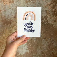 Load image into Gallery viewer, A white card with black text "you're my favorite" under a four stripe rainbow. The inner stripe is a thin light blue line. The next stripe is a thicker salmon colored line filled with darker orange dots. The third line is a thin black line. The outer line is a thicker salmon colored line with a darker orange line through it. The card is held in front of a brown rustic wall by a white woman.