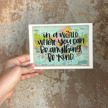 Load image into Gallery viewer, A blue, green, yellow, and pink flat map of the world with text "in a world where you can be anything, be kind" in black lettering over the map print. The map is held by a white woman, in front of a brown rustic wall.