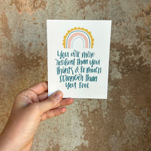 Load image into Gallery viewer, A white card with blue-green text "You are more resilient than you think & so much stronger than you feel" below a four stripe rainbow. The inner stripe is a thin light blue line of dots. The next stripe is a thicker salmon colored line. The third stripe is a thin blue line. The outer stripe is a thicker scalloped golden line. The card is held in front of a brown rustic wall by a white woman.