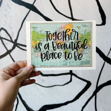 Load image into Gallery viewer, A blue, green, yellow, and pink flat map of the world with text "together is a beautiful place to be" in black lettering over the map print. The map is held in front of a white backdrop with spiraling black lines by a white woman.