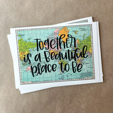 Load image into Gallery viewer, A stack of blue, green, yellow, and pink flat maps of the world with text "together is a beautiful place to be" in black lettering over the map print. The maps are lying on a large piece of brown paper.