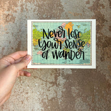 Load image into Gallery viewer, A blue, green, yellow, and pink flat map of the world with text "never lose your sense of wonder" in black lettering over the map print. The map is held in front of a brown rustic wall by a white woman.