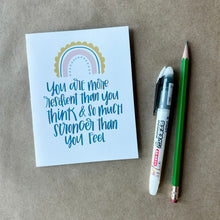 Load image into Gallery viewer, A white card with blue-green text "You are more resilient than you think & so much stronger than you feel" below a four stripe rainbow. The inner stripe is a thin light blue line of dots. The next stripe is a thicker salmon colored line. The third stripe is a thin blue line. The outer stripe is a thicker scalloped golden line. The card is lying on a piece of brown paper beside a pencil and a pen.