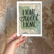 Load image into Gallery viewer, A vintage style map design of the state of Georgia with black lettered text "Home Sweet Home" over the print. The map is held in front of a brown rustic wall  by a white woman.