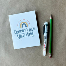 Load image into Gallery viewer, A white card with gray text "Sending love your way" under a four stripe rainbow. The inner stripe is a dark gray line. The next stripe is a golden line. The third stripe is a lighter gray line. The outer stripe is a line of red dots. The card is lying on a piece of brown paper beside a pencil and a pen.