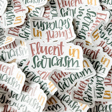Load image into Gallery viewer, A sticker with text "Fluent in Sarcasm" rests on a pile of the same sticker. "Fluent" is red, "in" is yellow, and "sarcasm" is dark green. The background color is white.