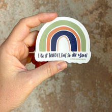 Load image into Gallery viewer, Dark blue text "Life is tough, but so are you!" under a four stripe rainbow. The inner stripe is thin and light pink, the next stripe is thick and dark blue, the third stripe is slightly less thick and salmon colored, and the last stripe is thicker and green. The background color is white. The sticker is held in front of a brown rustic wall by a white woman.