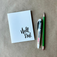 Load image into Gallery viewer, A white card with black text "Well, shit." in the bottom right corner. The card is lying on a piece of brown paper beside a pencil and a pen.