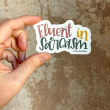 Load image into Gallery viewer, A white woman holds up a sticker with text "Fluent in Sarcasm" in front of a brown rustic wall. "Fluent" is red, "in" is yellow, and "sarcasm" is dark green. The background color is white.
