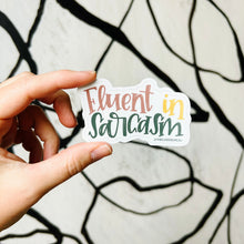 Load image into Gallery viewer, A white woman holds up a sticker with text "Fluent in Sarcasm" in front of a white background with spiraling streaks of black. "Fluent" is red, "in" is yellow, and "sarcasm" is dark green. The background color is white.
