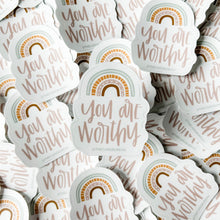 Load image into Gallery viewer, Light pink text "you are worthy" under a 4 stripe rainbow. The inner stripe is a golden brown. The next stripe is light pink with white arrow tip designs. The third stripe is a dashed golden line. The outer stripe is a blue-green line with a thin white line in the middle of it. The sticker is in front of a pile of the same sticker.