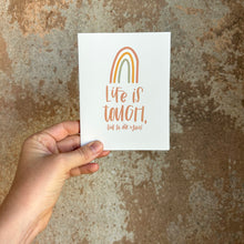 Load image into Gallery viewer, A white card with orange text "Life is tough, but so are you!" under a three stripe rainbow. The inner stripe of the rainbow is a green line, the middle stripe is an orange line, and the outer stripe is a red line. The card is held in front of a brown rustic wall by a white woman.
