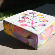 Load image into Gallery viewer, A closeup of a colorful abstract painting with colors pink, purple, yellow, orange, blue, and black painted over with white to show a plant in the negative colored space. The painting is laying on a tan canvas outdoors.