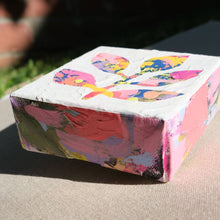 Load image into Gallery viewer, A closeup of a colorful abstract painting with colors pink, purple, yellow, orange, blue, and black painted over with white to show a stem with leaves in the negative colored space. The painting is laying on a tan canvas outdoors.