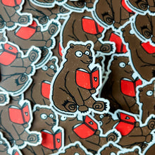 Load image into Gallery viewer, A sticker of a brown bear reading a red book with a heart on it with a white background color, in front of a pile of the same sticker.