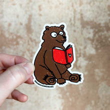 Load image into Gallery viewer, A white woman holds up a sticker of a brown bear reading a red book with a heart in front of a brown rustic wall.