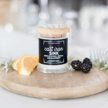 Load image into Gallery viewer, 7th Street Salvage's Cast Iron Sink candle sitting on a wooden cutting board with orange slices and black berries on the side. The candle label is black with white typography and the container has a wooden lid.