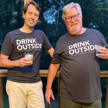 Load image into Gallery viewer, Two white men holding drinks with grey tees with "drink outside" printed in the center. They are leaning on a wooden rail with greenery in the background