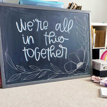 Load image into Gallery viewer, Larger Than Life's monthly chalkboard design. This month's board says "we're all in this together" in white lettering with a blue drop shadow. In the bottom right corner there is a flower with leaves.