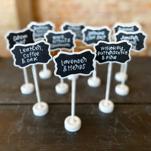 Load image into Gallery viewer, Small white stands with lettering. Each sign has different candle notes, like leather, coffee, and oak or lavender and herbs written in white.
