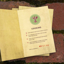 Load image into Gallery viewer, Yellow card open with green circle emblem of utensils. "Cooking" text in middle with checkboxes under and date with a stamp at the bottom. Brick background with moss in cracks