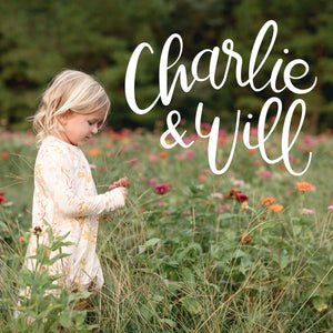 Handlettered white Charlie & Will logo on a photo of a young girl standing in a zinna field of flowers wearing a Charlie and Will dress. 
