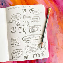 Load image into Gallery viewer, Photo of a sketch book of hand lettering designs of "macon sense" with pencil resting on the page and warm colored watercolor background.