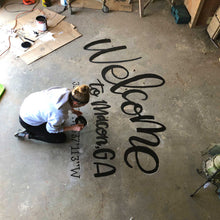 Load image into Gallery viewer, Mama Hawk Draws kneeling on concrete floor painting "welcome to macon, GA" with coordinates in black lettering