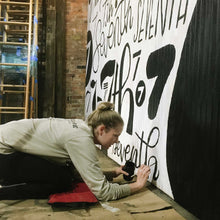 Load image into Gallery viewer, Mama Hawk Draws painting black lettering on the white wall of the 7th street salvage mural