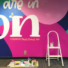 Load image into Gallery viewer, Closeup of the right of the macon airport mural with a ladder and a can of paint resting on it as well as some cans of paints and paintbrushes resting on a tarp on the ground to the left